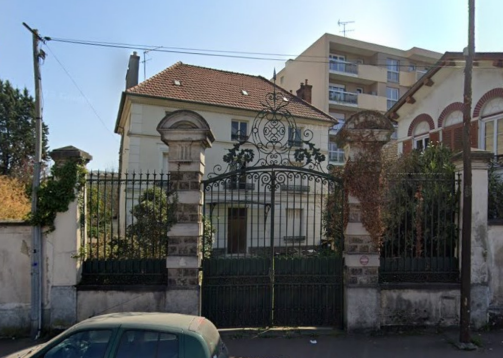 Façade du pavillon Dionnet à Livry-Gargan en 2023, derrière une grille en fer forgé, avec un portail monumental encadré de piliers en pierre.